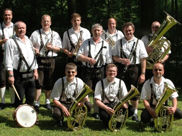 Elf Musiker in Tracht stehen mit Blasinstrumenten und einer Trommel auf einer Wiese.