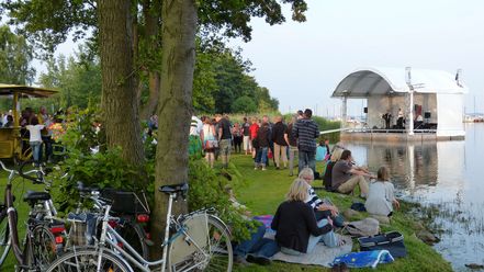 Menschen genießen ein Konzert auf einer Seebühne vor dem Fischerstübchen an einem sonnigen Tag.