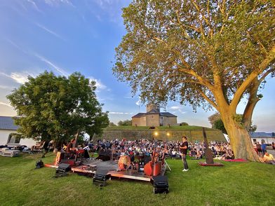 Open-Air-Konzert auf einer grünen Wiese vor einer historischen Festung bei Sonnenuntergang.