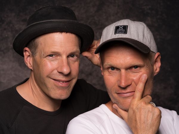 Two smiling men in casual clothes and hats pose in front of a dark background.