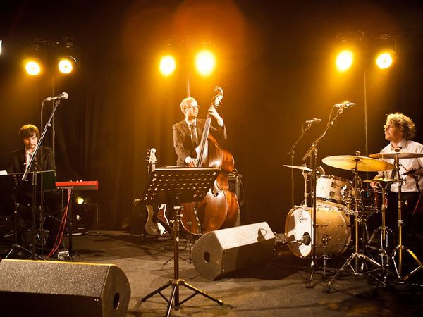 Jazzband mit Kontrabass, Schlagzeug und Klavier auf einer Bühne im warmen Scheinwerferlicht.